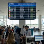 Suasana ruang tunggu keberangkatan di bandara dengan layar jadwal penerbangan besar dan pesawat di latar belakang.